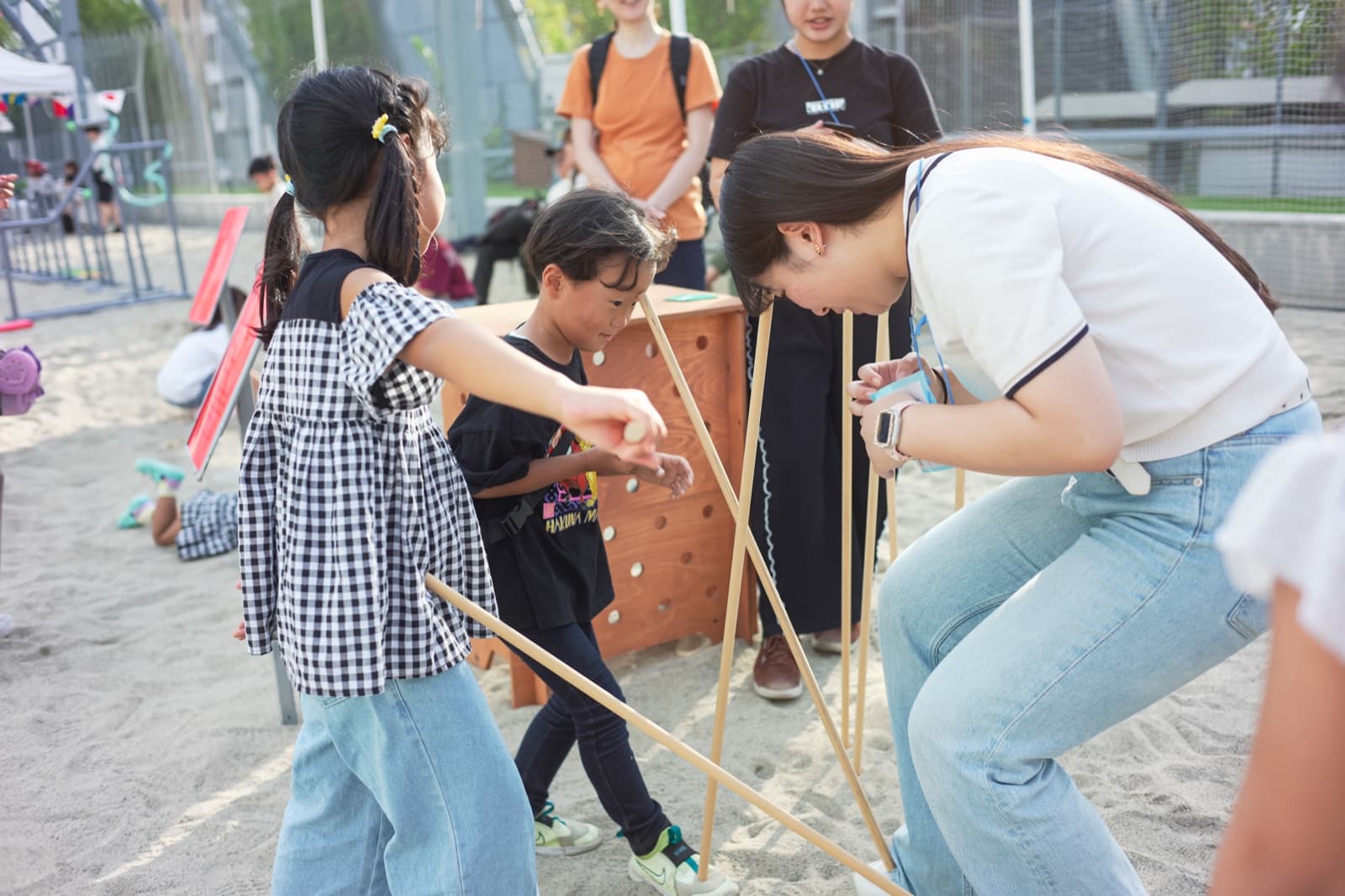 砂地の広場で、子どもと大人が長い棒を腕や頭で支え合いながら遊んでいる。