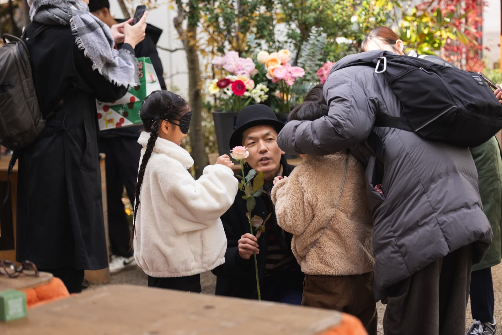 街角の花屋の前で、アイマスクをした子どもと大人が一輪の花を手に取り、香りを確かめている。