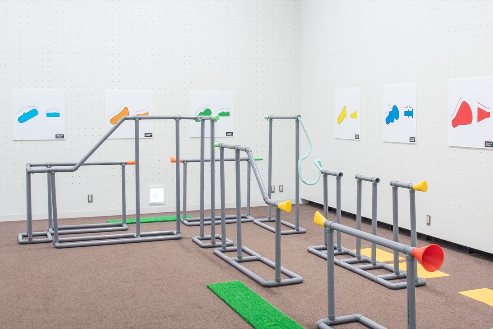 白い壁の部屋に、灰色の手すりパイプでつくられた迷路のようなコースが組まれている。足元には人工芝、壁には色とりどりの図形が貼られている。
